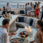 People socializing at an elegant outdoor party by the ocean, with food, drinks, and floral centerpieces on tables.