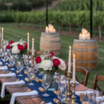 An elegantly set outdoor dining table with candles, flowers, and wine glasses under string lights, with vineyards in the background.