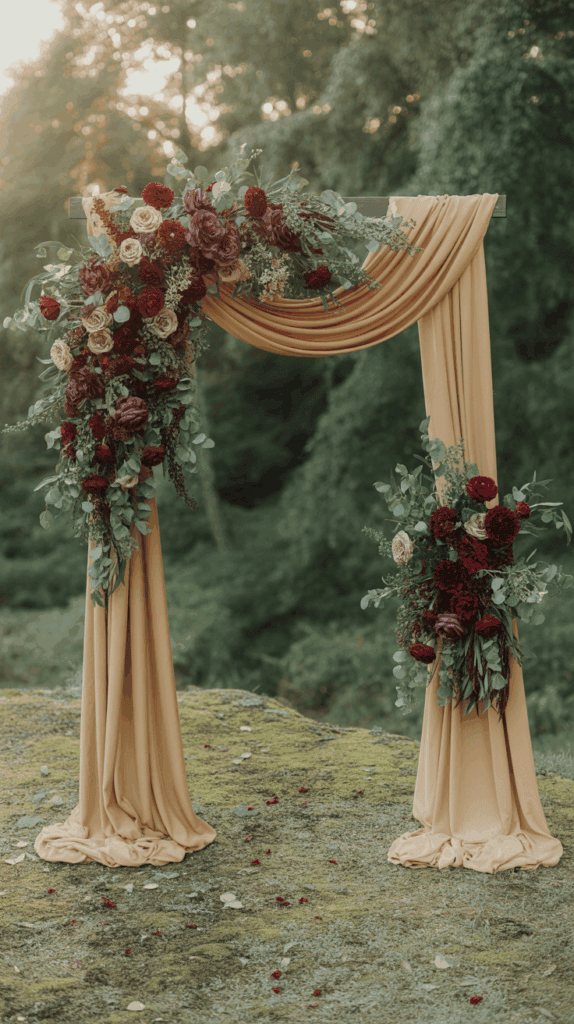 A decorative wedding arch adorned with flowing mustard-colored fabric and rich floral arrangements of red and mauve flowers, set against a lush green outdoor background.