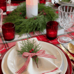 A festive table setting featuring a red and green checkered tablecloth, white plates with red edges, gold cutlery, and crystal glassware. A white candle surrounded by greenery forms the centerpiece, while a neatly folded white napkin with red trim rests on the plate, adorned with a small sprig of evergreen and tied with a red and white ribbon.