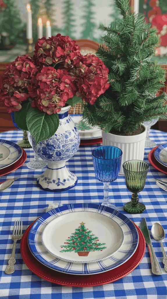A table set with a blue and white checkered tablecloth, featuring a festive place setting with plates, cutlery, and colorful glassware. A vase with vibrant red hydrangeas and a small decorative evergreen tree are displayed in the center.