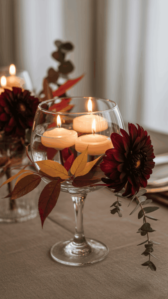 A glass goblet filled with water holds floating candles, decorated with red leaves and a dark red flower on a table.