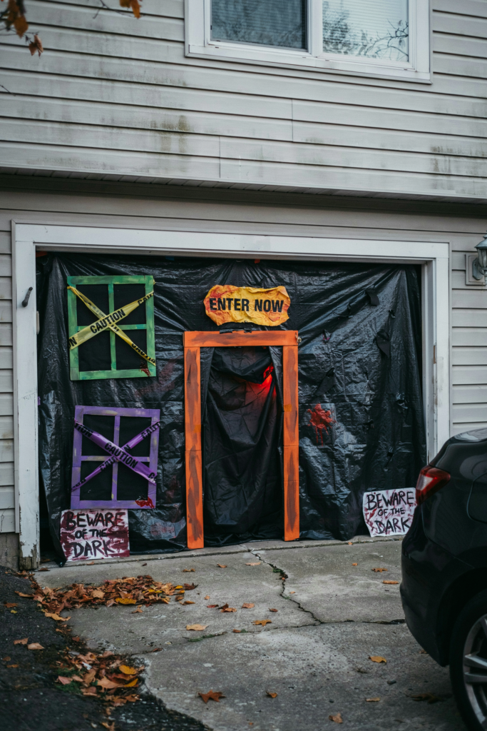 Garage door decorated for Halloween with black plastic, a faux orange doorway, and signs saying "Enter Now" and "Beware of the Dark."