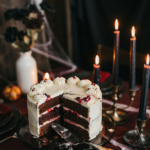 A sliced chocolate cake with white frosting and berry filling on a dark table with lit candles and cobwebs.