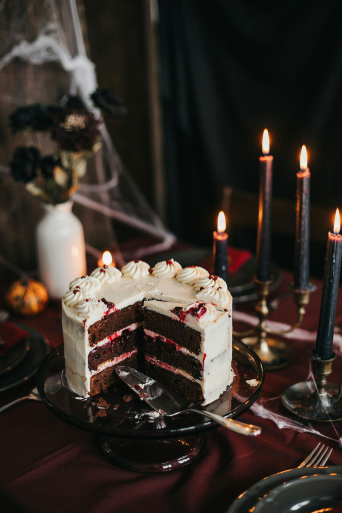 A sliced chocolate cake with white frosting and berry filling on a dark table with lit candles and cobwebs.