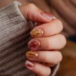 A hand with autumn-themed nail art, featuring red and yellow leaves with gold glitter accents, resting on a wooden surface.