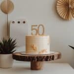 A white cake with "50" on top, decorated with golden accents, placed on a wooden stand surrounded by potted plants.