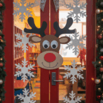A decorated red double door with large snowflake decals and a reindeer face on the glass. A silver bow is above the doors, and the frame is adorned with a garland and ornaments. The interior is visible through the glass, showing a colorful room with bookshelves.