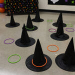 Ring toss game set up with witch hats as targets, surrounded by colorful rings; a Halloween-themed party backdrop is visible.