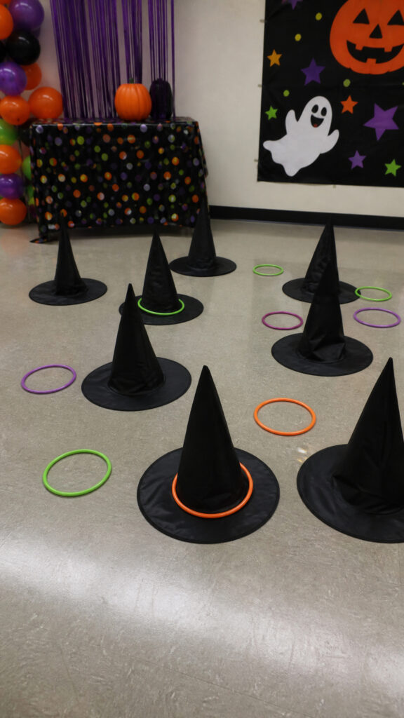 Ring toss game set up with witch hats as targets, surrounded by colorful rings; a Halloween-themed party backdrop is visible.