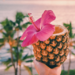 A hand holding a hollowed-out pineapple decorated with a pink hibiscus flower, with a blurred background of palm trees and a sunset over the ocean.