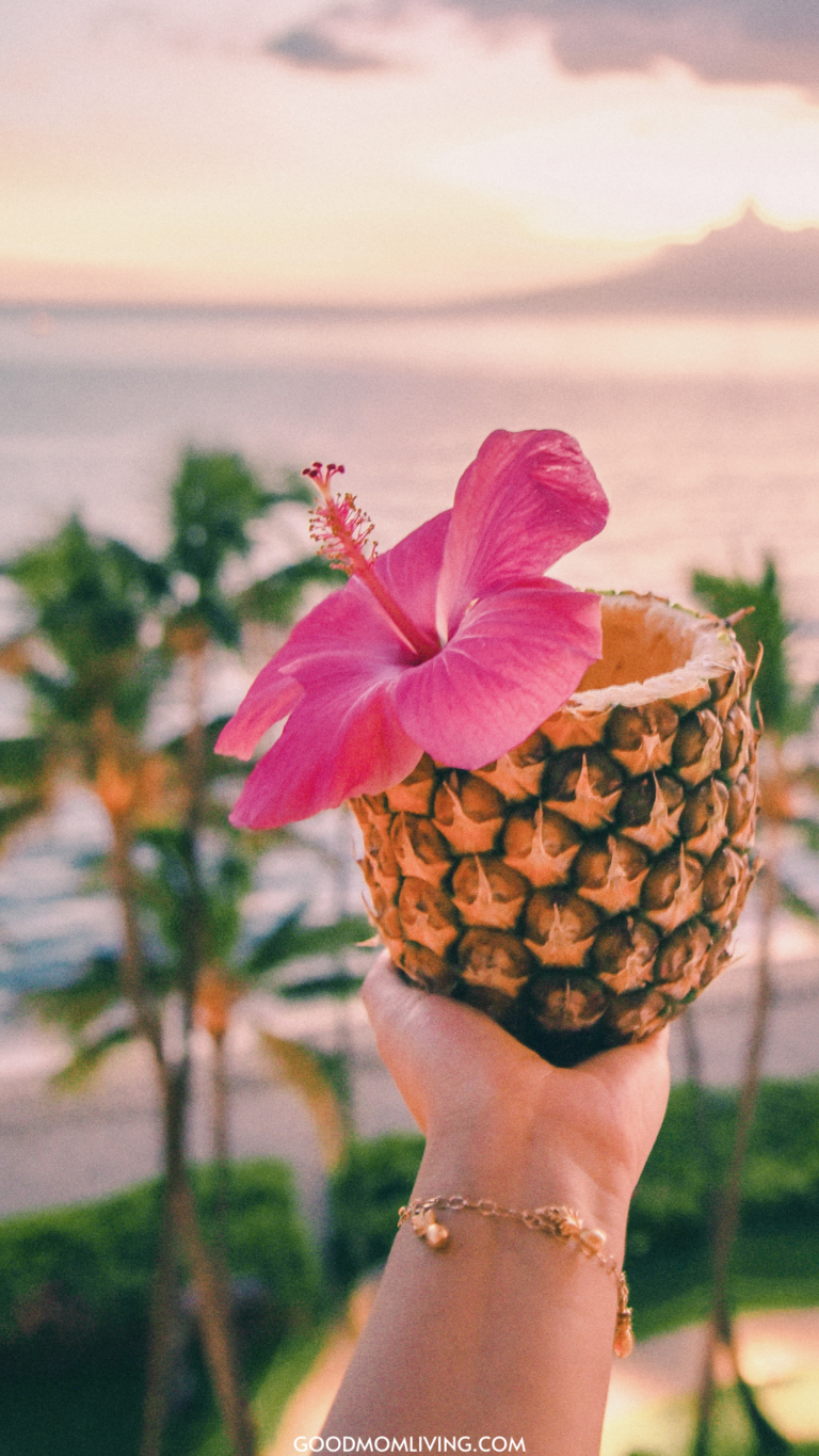 A hand holding a hollowed-out pineapple decorated with a pink hibiscus flower, with a blurred background of palm trees and a sunset over the ocean.