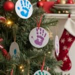 A Christmas tree decorated with round ornaments featuring colorful handprints, along with red and gold baubles. A red stocking and wrapped presents are visible in the background.