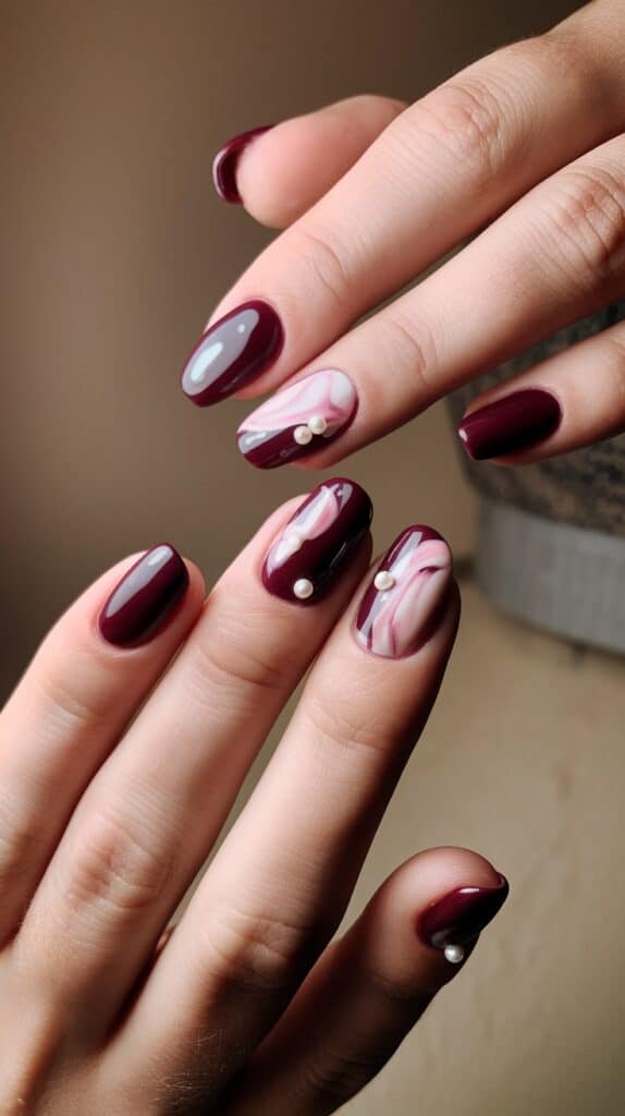 Close-up of hands displaying burgundy nails with a glossy finish, featuring marble designs with pearls on accent nails.