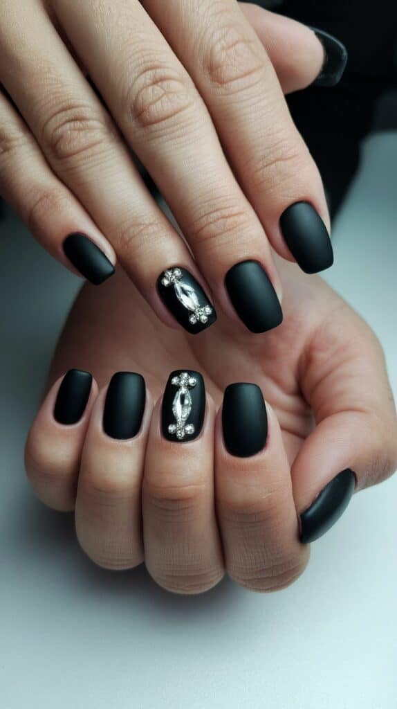 A hand with matte black nails, one featuring silver rhinestone embellishments.
