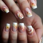Hands with nail art featuring autumn leaves in orange and yellow on a white background, with clear polish for a glossy finish.