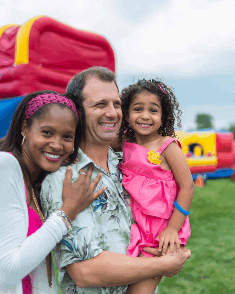 A smiling family of three poses outdoors near a colorful inflatable structure, with a woman in white and pink, a man in a floral shirt, and a young girl in a pink dress.