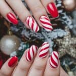 Hands with festively painted nails featuring a red and white candy cane stripe pattern against a holiday-themed background with ornaments.