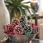 A decorative bowl filled with colorful, painted pine cones, sitting on a glass table in a stylish interior setting.