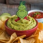 A bowl of guacamole topped with a Grinch decoration, surrounded by tortilla chips, with a small bowl of salsa in the background.