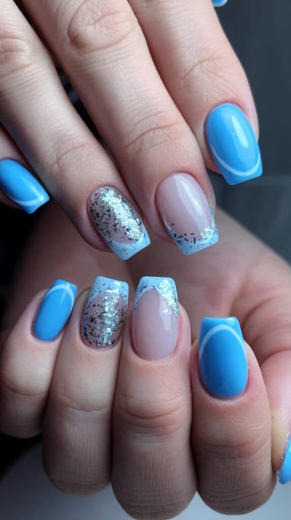 A hand with nails painted in a variety of light blue and glittery silver designs, alternating between solid blue, blue French tips, and silver glitter accents.