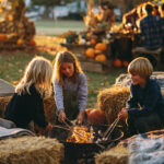 Three children roast marshmallows over a fire pit surrounded by hay bales, with pumpkins and autumn decorations in the background.