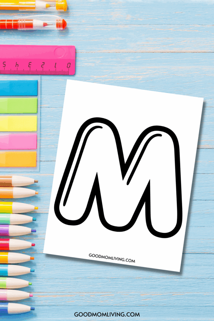 A large outlined letter 'M' on a piece of paper, surrounded by colorful pens, a pink ruler, and sticky notes on a blue wooden background.