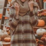 A person wearing a plaid shirt and knitted scarf stands in an autumn-themed setting with pumpkins and a ladder adorned with fall leaves in the background.