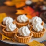 Plate of mini tarts topped with whipped cream, surrounded by colorful autumn leaves on a wooden table.