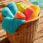 A wicker basket filled with brightly colored towels sits on a wooden deck beside a pool.