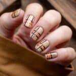 A close-up of a hand with plaid-themed nail art featuring beige, orange, and dark brown stripes.