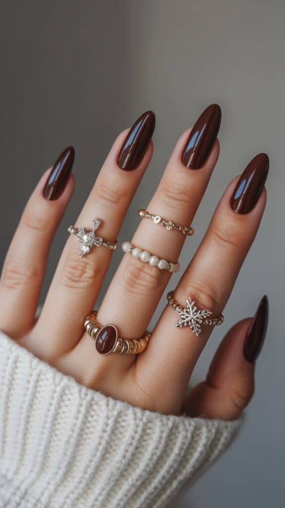 A hand with long, almond-shaped, dark chocolate brown nails adorned with multiple rings, including a pearl ring and a decorative snowflake ring, against a blurred background.