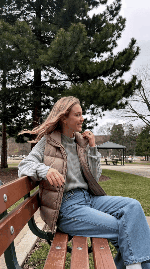 A person with long hair wearing a grey sweater and brown puffer vest sits on a park bench, smiling, with trees and a gazebo in the background.